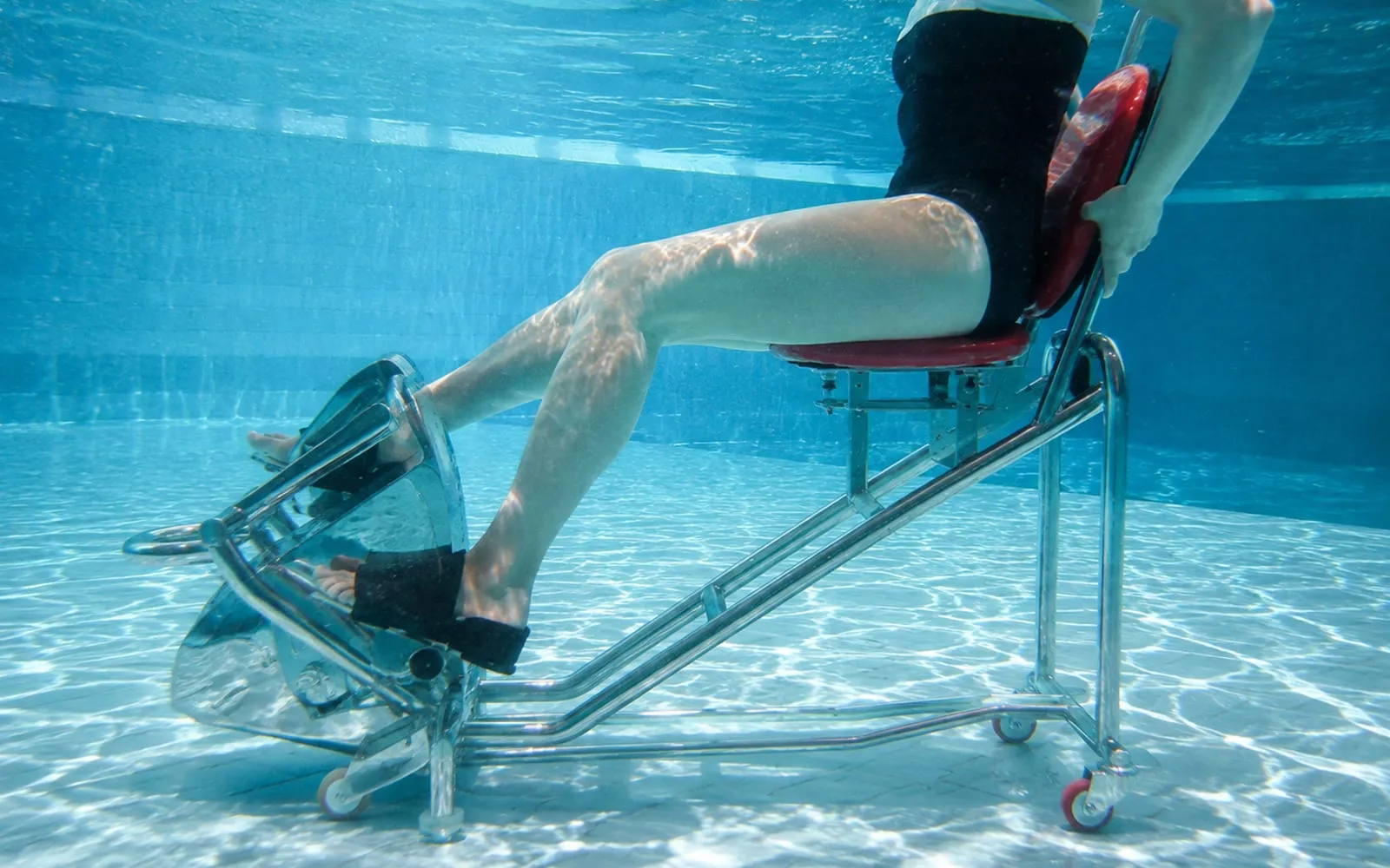 Unter Wasser: ein Trainingsgerät mit Sitz und Pedalen am Beckenboden, eine Person tritt in die Pedale.