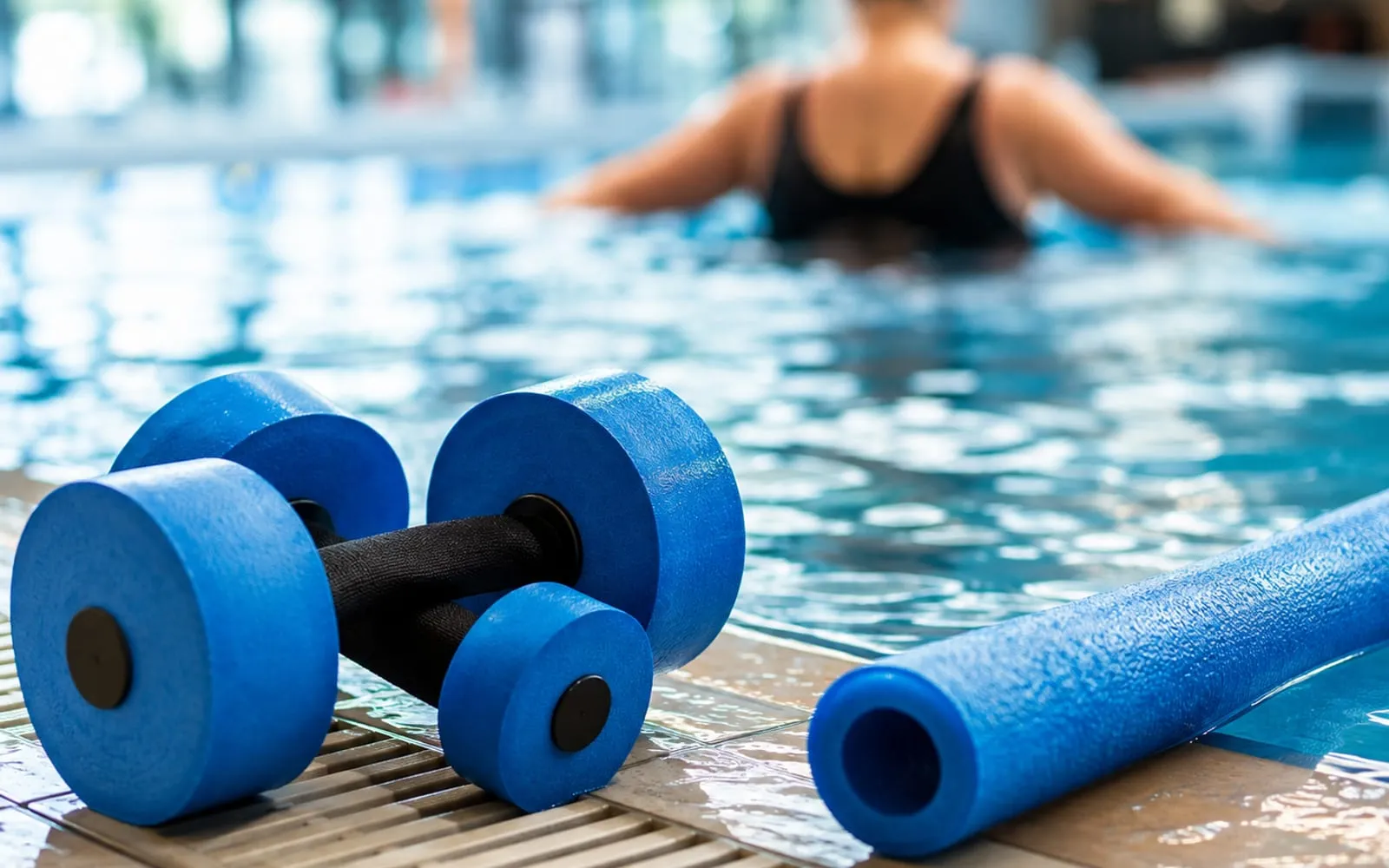Zwei blaue Schaumstoffhanteln und eine Poolnudel liegen am Beckenrand, dahinter unscharf das Wasser.