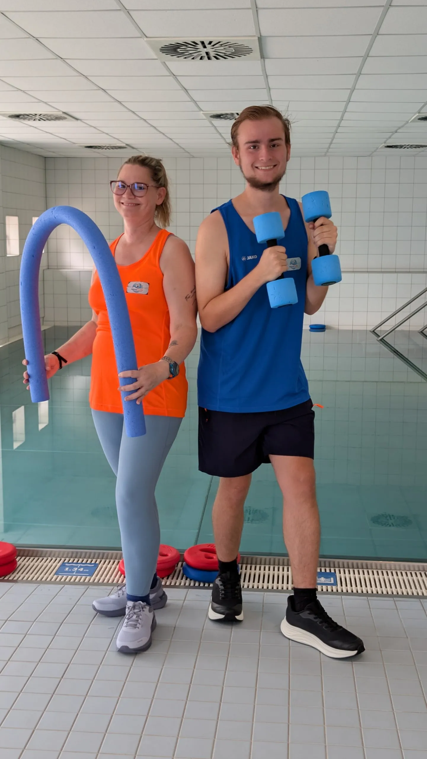 Nicole und Colin Gulbinski nebeneinander am Beckenrand eines Hallenbads: Nicole im orangefarbenen Aquaflow-Shirt mit einer blauen Poolnudel, Colin im blauen Aquaflow-Shirt mit zwei Aqua-Hanteln, beide lächeln in die Kamera.